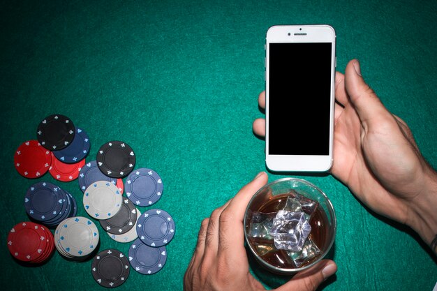 Poker player's hand showing mobile phone and holding whiskey glass on poker table