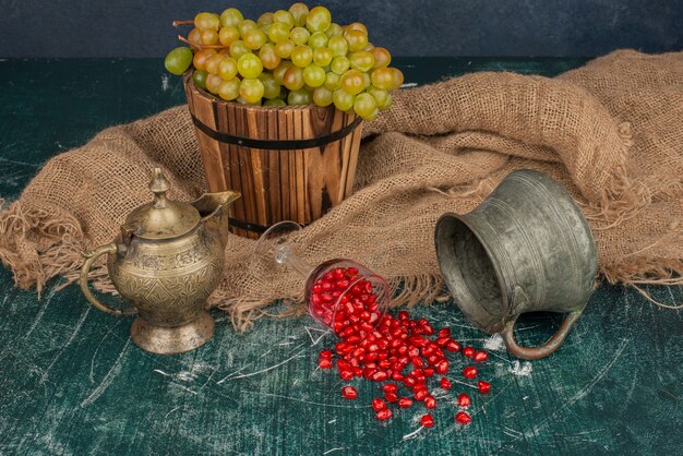 Pomegranate seeds and bucket of grapes on marble table with vase and teapot