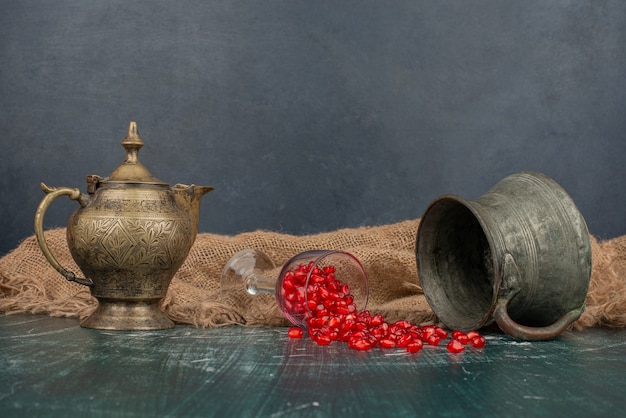 Pomegranate seeds scattered on marble table with vase and teapot.