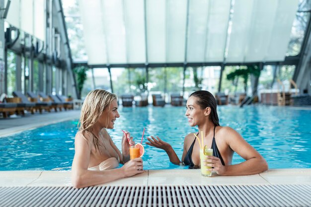 健康的な飲み物を飲みながらリラックスするプールサイドの友人スパスイミングプールスパ屋内プールでリラックスする官能的な若い女性