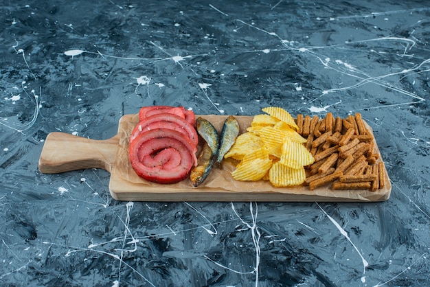 Сало свинины, рыба, чипсы и панировочные сухари на разделочной доске, на синем фоне.