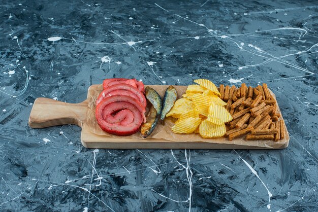 Сало свинины, рыба, чипсы и панировочные сухари на разделочной доске, на синем фоне.