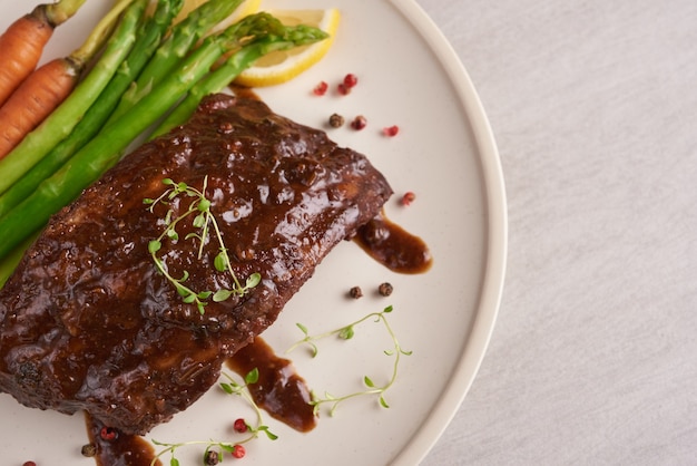 Pork roasted, grilled spare ribs from a summer BBQ served with Vegetables, asparagus, baby carrots, fresh tomatoes and  spices. smoked ribs in white plate on stone surface . top view.