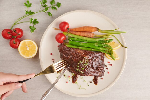 Pork roasted, grilled spare ribs from a summer BBQ served with vegetables, asparagus, baby carrots, fresh tomatoes, spices in white plate. woman hands with fork and knife eating spare ribs. top view.