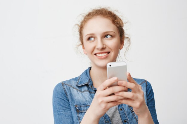 Portait of cute tender redhead teenage girl with messy hair, looking aside and smiling while holding smartphone