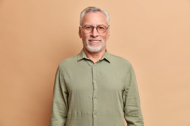 Portait of handsome European elderly man smiles positively enjoys retirement wears shirt and spectacles has perfect white teeth isolated over beige studio wall
