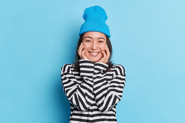 Portait of pleasant looking cheerful Asian woman keeps hands on cheeks smiles gently wears striped jumper stylish hat expresses sincere emotions 