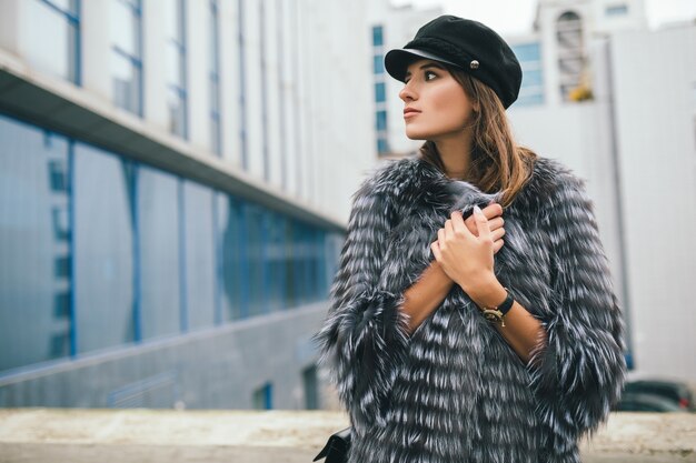 Portrair of fashionable woman walking in city in warm fur coat wearing black cap