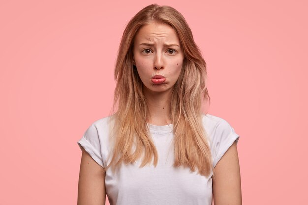 Portrait of abused blonde woman sulks and purses lips, being insulted by someone and waits for apologies, has straight hair, isolated over pink wall