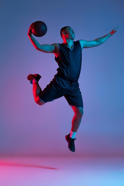 Portrait of active boy basketball player in a jump training isolated over gradient purple pink background