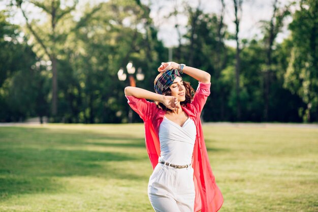 Portrait of active brunette girl in bandana posing in summer park. She wears white clothes, long pink shirt. She is smiling with closed eyes.