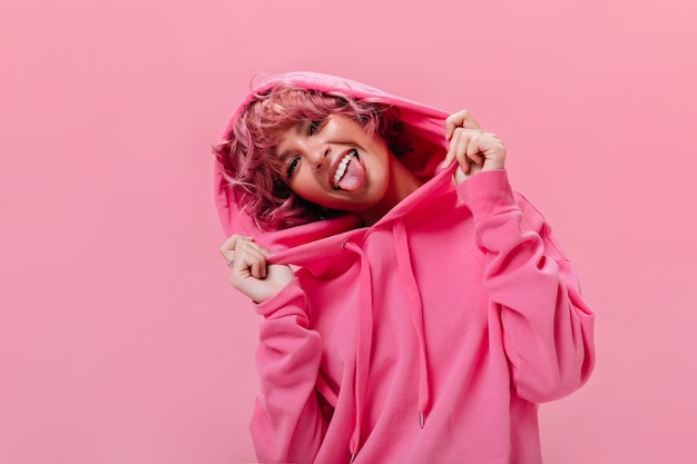 Portrait of active cheerful pink-haired woman in fuchsia oversized hoodie shows tongue and makes funny face on isolated wall 