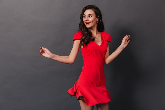 Portrait of active girl in red dress dancing with smile against black background. Brunette in massive earrings has fun.