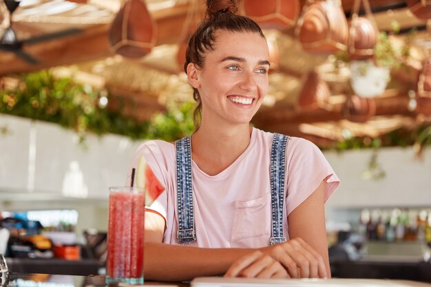 Ritratto di adorabile modello femminile dagli occhi blu con nodo di capelli indossa una tuta, gode di bere frullati di frutta, guarda felicemente da qualche parte mentre si siede in un accogliente ristorante. bella ragazza adolescente nella caffetteria