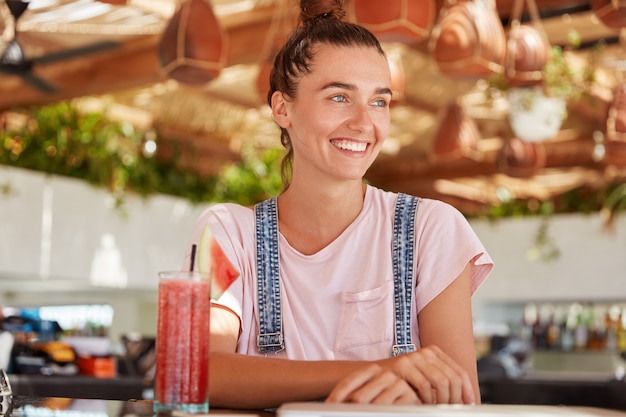 Portrait of adorable blue eyed female model with hair knot wears overalls, enjoys drinking fruit smoothie, looks happily somewhere while sits in cozy restaurant. Beautiful teenage girl in coffee shop