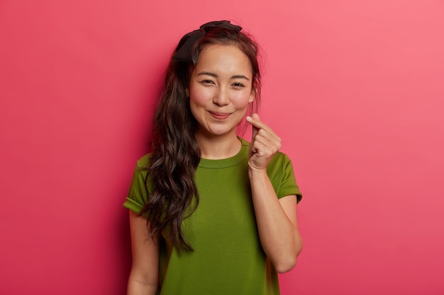 愛らしいブルネットの少女の肖像画は愛と幸福を広め、ハートのサイン、指で愛情の韓国のシンボルを示し、明るいピンクの背景の上に分離された緑のTシャツを着ています