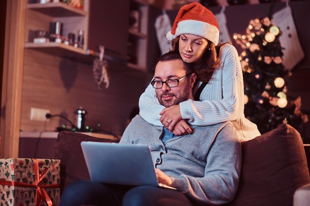 사랑스러운 커플의 초상화 - 산타 모자를 쓴 매력적인 여성이 남자를 껴안고 노트북을 사용합니다. 크리스마스 이브를 축하하는 여성.