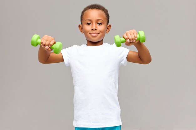 Portrait of adorable fit athletic dark skinned boy in white blank t-shirt doing morning physical exercise routine for biceps using two green dumbbells having energetic happy facial expression