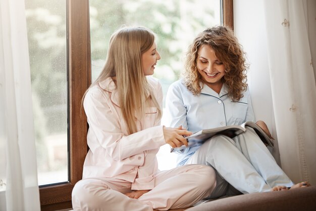 Portrait of adorable friends sitting on window sill in pyjamas, reading magazine