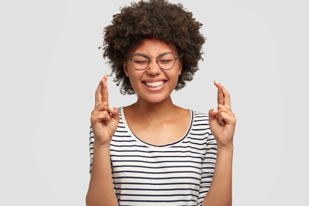 Portrait of adorable young female with positive smile, has Afro hairstyle and dark skin, crosses fingers as believes in good luck, dressed in striped sweater, isolated over white wall