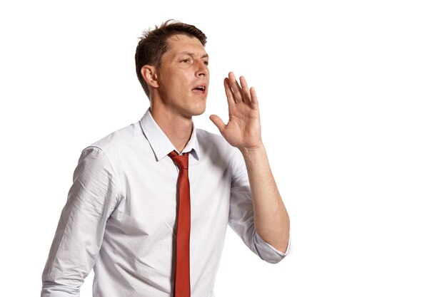 Portrait of an adult brunet male with brown eyes, wearing in a white shirt and a red tie. He is acting like calling someone while posing in a studio isolated over a white background. Concept of gestic