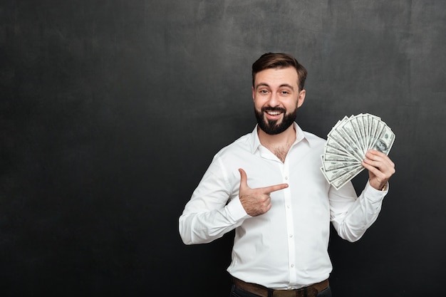 Portrait of adult man in white shirt posing on camera with fan of 100 dollar bills in hand, being rich and happy over dark gray