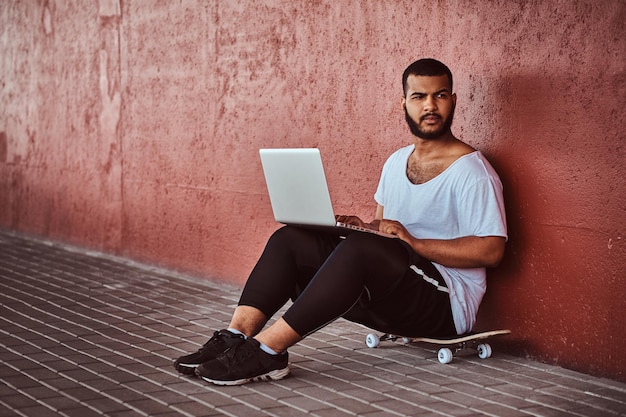 Foto gratuita il ritratto di un ragazzo barbuto afroamericano vestito con una camicia bianca e pantaloncini sportivi tiene su un laptop mentre è seduto su uno skateboard sotto il ponte, guardando lontano.