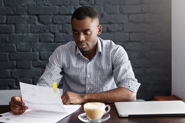 Portrait of African-American businessman in checkered shirt examining closely piece of paper in his hands