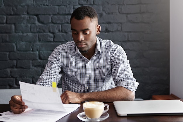 Portrait of African-American businessman in checkered shirt examining closely piece of paper in his hands