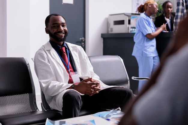 Портрет афроамериканского врача в вестибюле, сидящего на местах в зале ожидания перед медицинской консультацией с пациентами. Врач общей практики работает над здравоохранением в центре.