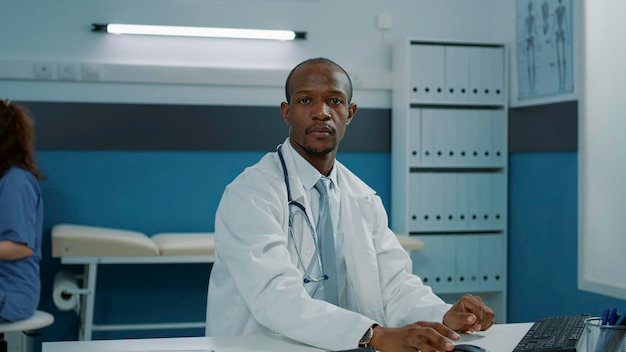 Портрет афроамериканского врача, использующего компьютер в кабинете, работающего над поддержкой здравоохранения. Врач общей практики в белом халате и со стетоскопом, уверенный в своих услугах и опыте.