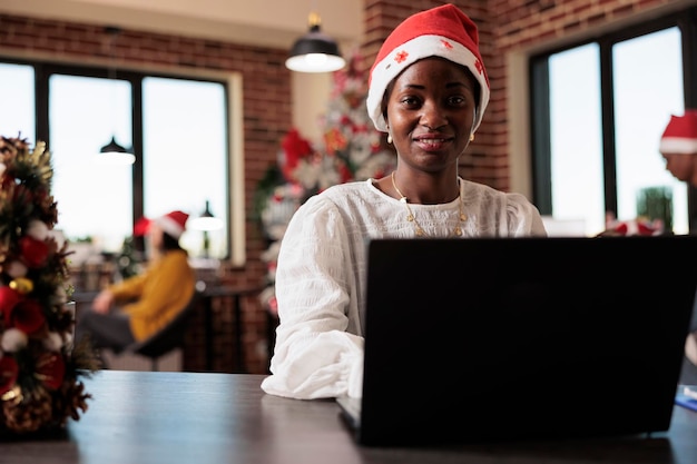 Портрет афроамериканского сотрудника в шляпе Санты, работающего по делам в офисе компании, наполненном рождественскими украшениями и елочными огнями. Использование ноутбука на стартовом рабочем месте с рождественским декором.