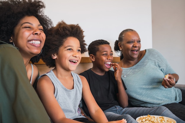 Portrait of African American grandmother, mother and children watching a movie and eating popcorn while sitting on sofa at home. Family and lifestyle concept.