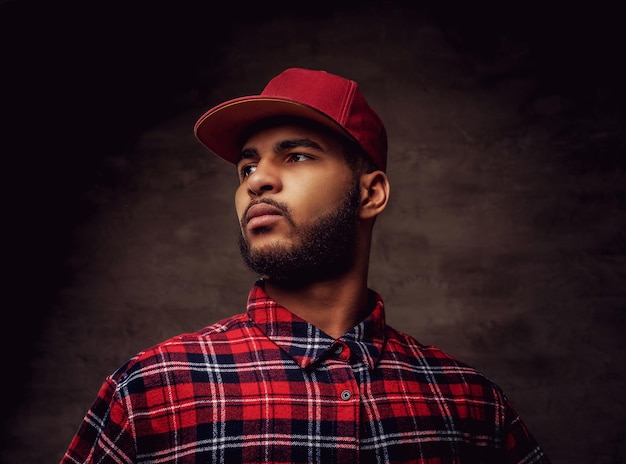 Portrait of an African-American hipster guy dressed in a red fleece shirt and cap at the studio. Isolated on a dark textured background.