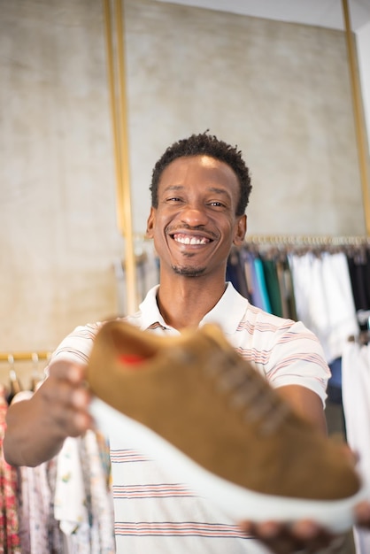 그의 신발 선택에 만족한 아프리카계 미국인 남자의 초상화. 부티크에서 새 신발을 들고 카메라에 보여주고 웃고 있는 행복한 검은 머리 남자. 남성복 및 의류 비즈니스 개념 구매