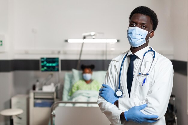 Portrait of african american physician doctor with protection face mask to prevent infection with covid19 looking into camera while standing in hospital ward. In background patient recovering