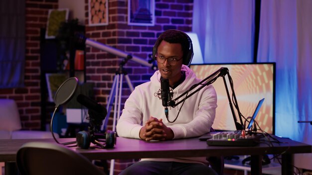 Portrait of african american podcaster adjusting digital audio mixer while talking live to audience using microphone. Male online radio host smiling confident at camera while recording in home studio.