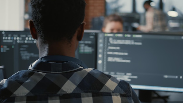 Portrait of african american programer online cloud computing on laptop sitting at desk with multiple screens parsing code. Over shoulder view of app developer working on user interface.