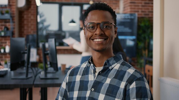 Portrait of african american programer sitting down working on laptop arranging glasses looking up and smiling at camera. System engineer using portable computer computing big data.