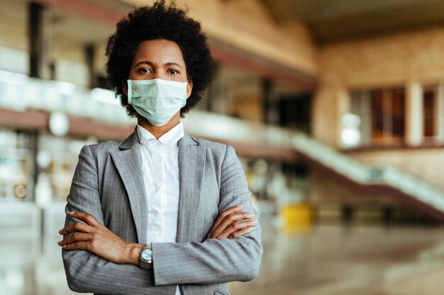 Портрет афроамериканки в защитной маске, стоящей со скрещенными руками в аэропорту во время эпидемии вируса