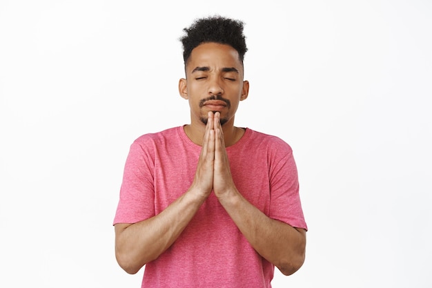 Portrait of african american young man praying, holding hands in supplicating, namaste gesture, close eyes, pleading god, standing in pink t-shirt against white background