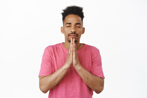 祈るアフリカ系アメリカ人の若い男の肖像画、嘆願、ナマステのジェスチャー、目を閉じて、神を懇願する、白い背景のピンクのTシャツに立って手をつないで