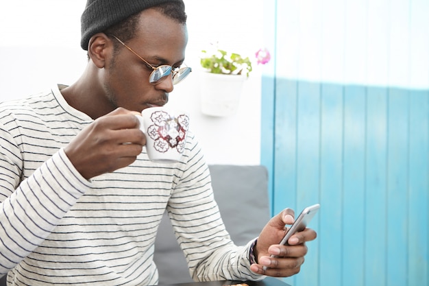 Ritratto di maschio africano in abiti eleganti con tazza, bere caffè fresco, navigare in internet e controllare newsfeed sui social media, utilizzando il telefono cellulare durante la colazione al bar con comodi posti a sedere