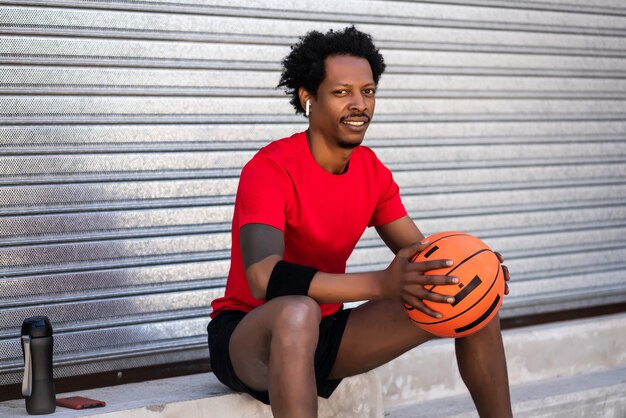 Портрет мужчины афро-спортсмена, держащего баскетбольный мяч и расслабляющегося после тренировки, сидя на открытом воздухе. Спорт и здоровый образ жизни.