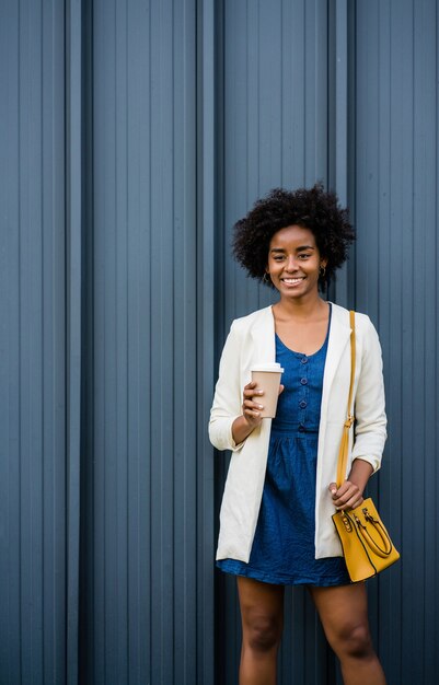 通りで屋外に立っている間コーヒーを保持しているアフロビジネス女性の肖像画。ビジネスと都市のコンセプト。