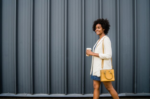 通りを屋外で歩きながらコーヒーを持っているアフロビジネス女性の肖像画。ビジネスと都市のコンセプト。