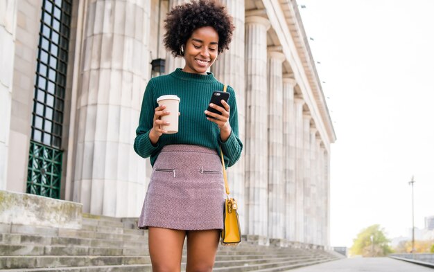 Портрет афро-деловой женщины, использующей свой мобильный телефон и держащей чашку кофе во время прогулки по улице