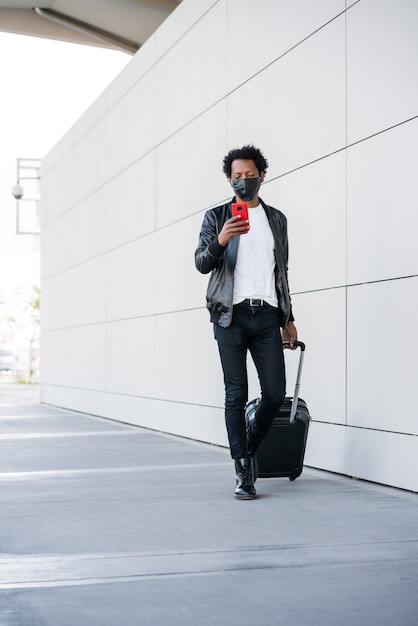 Portrait of afro tourist man typing on the phone and carrying suitcase while walking outdoors on the street