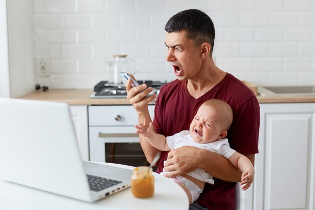 Portrait of aggressive man wearing maroon casual t shirt holding smart phone and screaming angrily, holding baby girl or boy, having problems at work while taking care of infant.