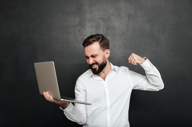 Portrait of agressive bearded man holding silver personal computer and throwing punch in screen, isolated over dark gray wall